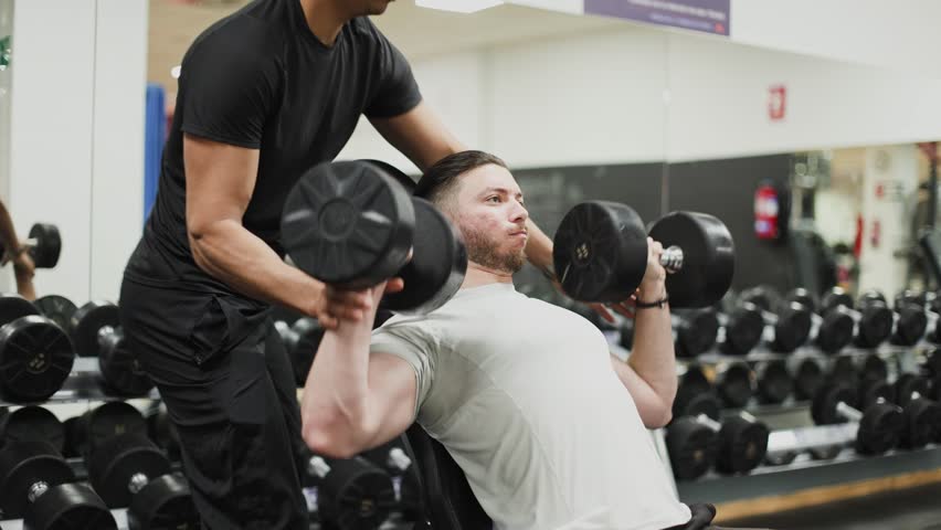 Dedicated personal trainer coaching and spotting a male athlete performing an intense incline dumbbell press for chest and shoulder development