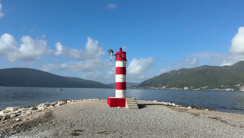 A red-and-white coastal lighthouse stands on a rocky shore under a clear blue sky, surrounded by calm sea and distant mountains. Perfect for travel, maritime, navigation and scenic landscape themes.