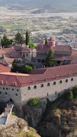 Vertical aerial forward view over a cliff-top monastery with the city of Meteora visible in the background, Greece.