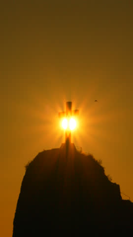 Cross, mountain, sunset, religious monument silhouetted against a golden sky with sunburst rays. Vertical video.