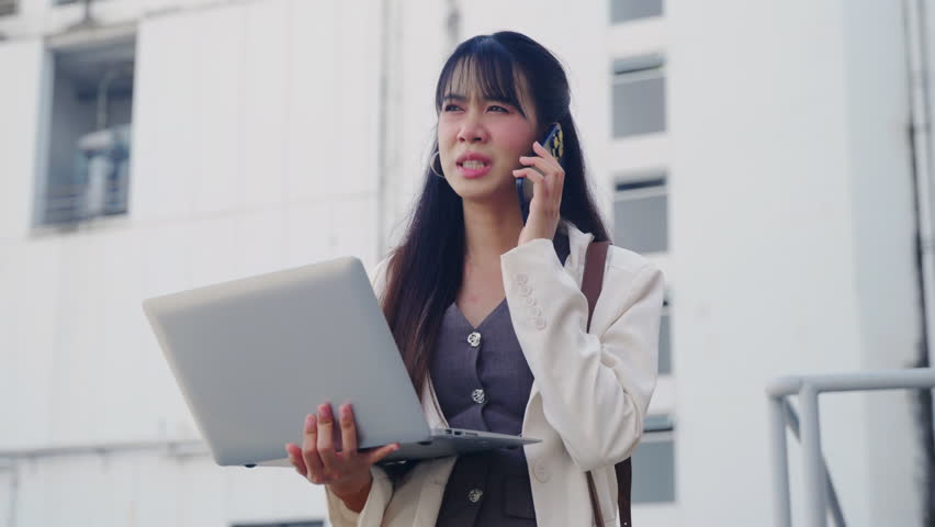 Young business businesswoman, traveling for work, appears stressed talking on phone, holding laptop, walking outdoor on bustling city street, modern office architecture visible.