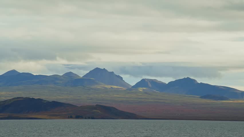 A wide shot of Eyjafjörður, a long fjord in Northern Iceland, with a tranquil body of water in the foreground and a range of majestic mountains under a cloudy sky in the background
