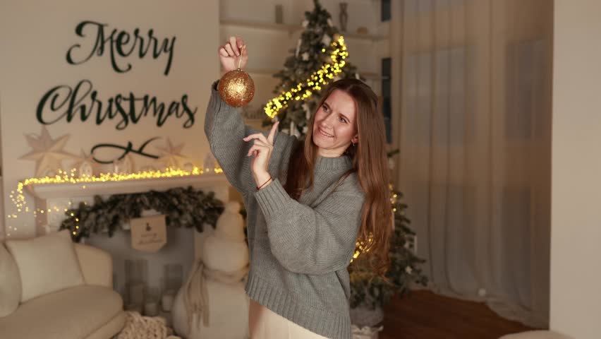 Cute woman in knitted sweater posing in hand toy balls to for Christmas tree in interior festive living room. Happy New Year. Cute girl holding Christmas ornament for decorate on Xmas tree at home
