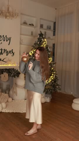 Cute woman in knitted sweater posing in hand toy balls to for Christmas tree in interior festive living room. Happy New Year. Cute girl holding Christmas ornament for decorate on Xmas tree at home