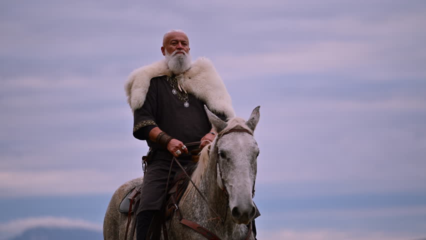 Elderly Viking warrior in fur cloak riding gray horse across open field at sunrise, mountains and distant cross behind him, moody cinematic Norse atmosphere cinematic slow motion 