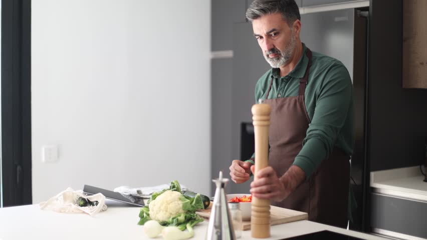 Confident middle aged chef wearing an apron preparing a healthy meal. He is seasoning a fresh vegetable salad with pepper, salt, and olive oil