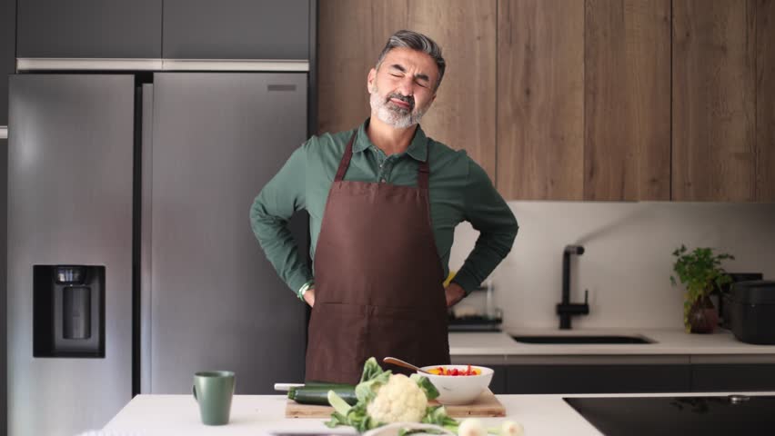 Tired mature man with a beard wearing an apron feeling backache and neck pain while preparing food. Uncomfortable senior suffering at home