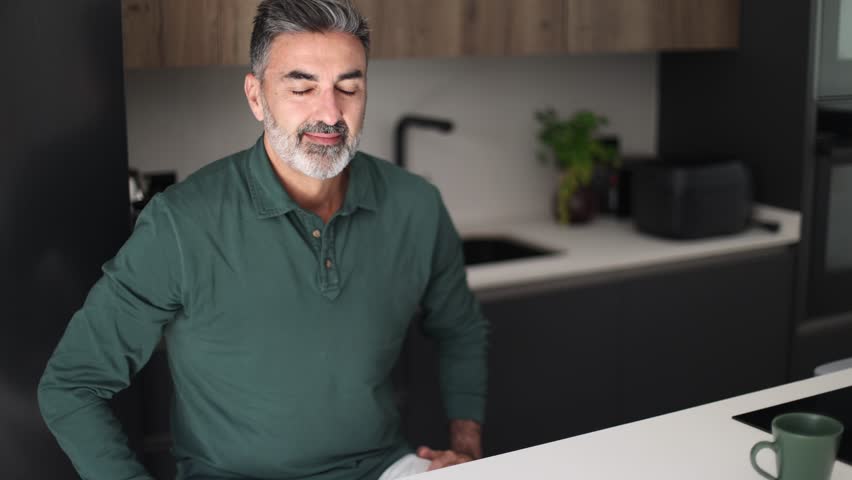 Relaxed mature man with a gray beard enjoying a tranquil moment at home. He takes a deep breath, smiles, and feels happy and peaceful