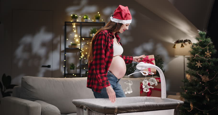 A pregnant woman wearing a Santa hat prepares a baby crib for the upcoming holidays. A pregnant woman wearing a Santa hat stands near a baby crib in a festive Christmas setting