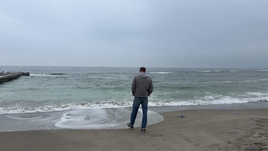 Man standing alone on sandy beach looking at rough sea. Overcast cold day Black Sea Odessa Ukraine. Contemplation, solitude, relax, rest during vacations.