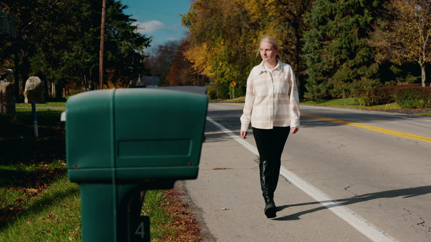 Woman walking toward a roadside mailbox to collect incoming mail on a sunny autumn day in a suburban neighborhood. High quality 4k footage