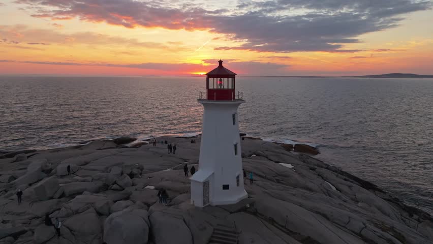 Cinematic Aerial View Of The Historic Peggys Cove Lighthouse In Halifax, Nova Scotia, Glowing In Warm Evening Light, With Its Reflection Appearing On Smooth Granite Surfaces And Rugged Atlantic Coast