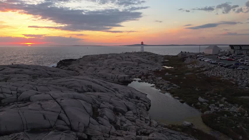 Cinematic Aerial View Peggys Cove Lighthouse In Halifax, Nova Scotia, Surrounded By Rugged Rock Formations, With Warm Sunset Light Casting A Golden Reflection Across The Granite Shore And Highlighting
