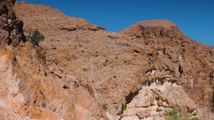 Footage of the rugged, dry mountains of Oman under a clear blue sky. The landscape is characterized by rocky terrain, brown and orange hues, and sparse vegetation, showcasing the country's unique natu
