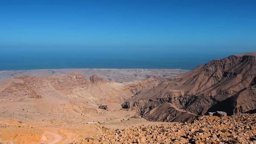A wide angle shot of a rugged mountain and desert landscape in Oman, with a clear blue sky over a vast expanse of water in the distance. The scene captures the arid beauty of the Middle Eastern terrai