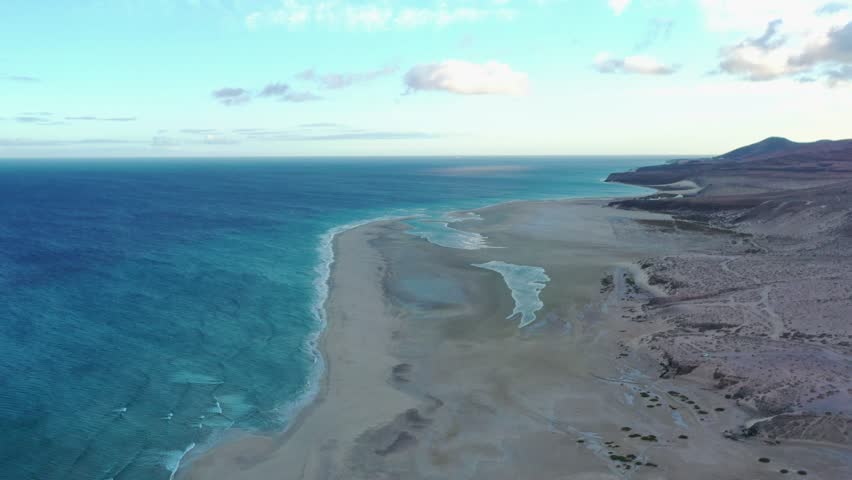 High-angle aerial shot of Fuerteventura’s iconic La Barca Beach at dusk, with glowing skies and gentle ocean textures.