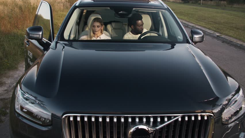 Couple seated inside dark luxury car preparing for drive. Man holding steering wheel adjusting controls. Woman fastening seatbelt beside him focused on buckling. Calm late afternoon on quiet roadside.