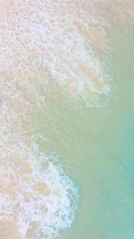 High-angle drone footage of a lone woman running through foamy surf along a bright turquoise tropical shoreline, trendy high quality clip good for destination branding, wellness edits, fitness vlogs