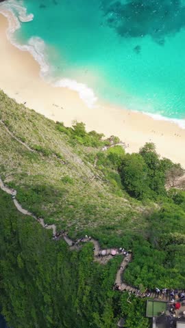 Aerial drone footage of Kelingking Beach in Bali showing steep hiking path, lush cliffs and turquoise waves. Ideal for travel vlogs, tourism promos, adventure themes and beach holiday online ads.