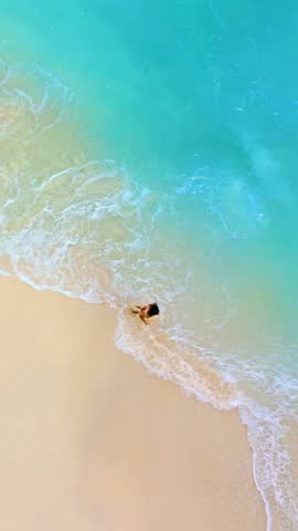 Aerial drone shot from above of a woman running along the shoreline where turquoise waves meet smooth tropical sand, ideal for travel promos, wellness retreats, freedom concepts, website headers and