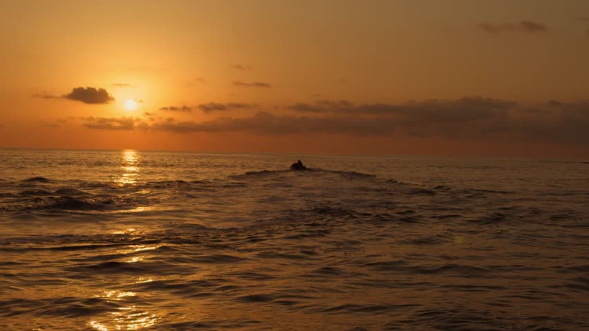 Jet ski rider carving sharply through golden sunset waves, creating a dramatic spray as warm light reflects across the ocean while another rider moves in the distance under an orange sky.