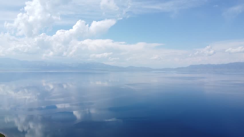 Experience the serene beauty of a calm lake reflecting the blue sky and clouds. An aerial view captures the tranquil water landscape with mountains in the distance.