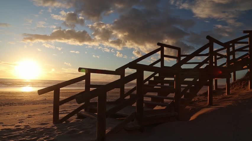 Lone traveler ascends wooden stairs on a Spanish beach, Cuesta Maneli, Huelva at sunset sunrise, silhouetted against the dramatic golden sky. Travel, peace, adventure.