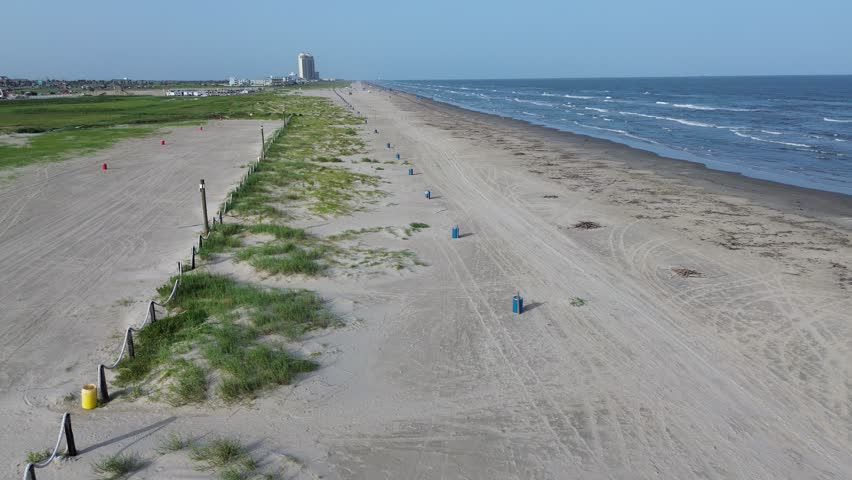 Expansive beach near Seawall Boulevard, Galveston, Texas with blue trash bins, grassy dunes, rope fencing, distant buildings, and tower structure, framed by rhythmic waves and clear Gulf horizon. USA