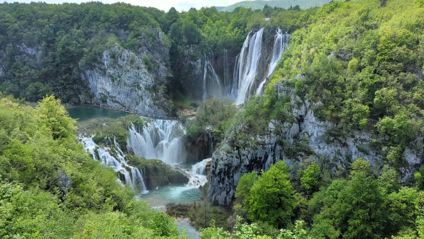 Stunning aerial view of cascading waterfalls flowing through lush green forest canyon with turquoise pools under dramatic summer sky in Plitvice Lakes National Park Croatia