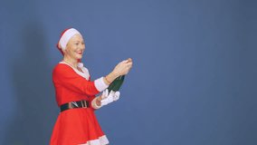 Happy senior woman in a mrs. Claus outfit celebrating christmas and new year by excitedly popping a bottle of sparkling wine on blue background - Powered by Shutterstock - Get 15% off with code: PIKWIZARD15