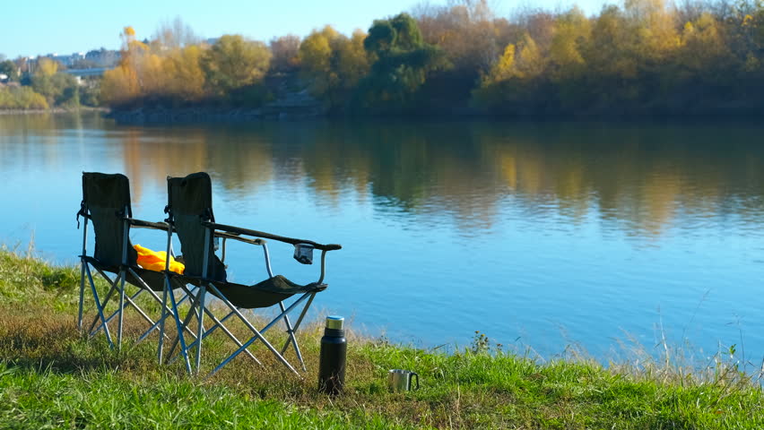 Two empty camping chairs by a peaceful river on a sunny day. Serene landscape with two empty folding chairs and a thermos on the green riverbank, offering a perfect spot for relaxation, a picnic