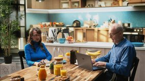 Elderly husband and wife enjoying morning routine at breakfast table, serving coffee together and navigating the web on laptop. Joyous bonding and natural lifestyle during retirement. Camera A. - Powered by Shutterstock - Get 15% off with code: PIKWIZARD15