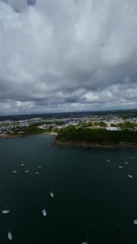 Aerial drone view France Atlantic peninsula charm, Brittany coastal town residential streets, sports field and boats in calm harbor under cloudy sky.