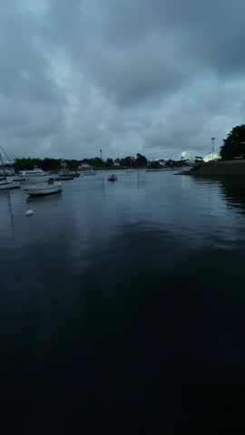 FPV drone flies fast across France Atlantic marina, Brittany harbor fills with colorful boats in calm water beside coastal homes under overcast sky.