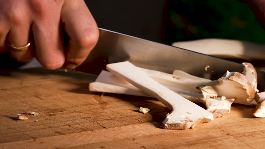 Close-up of king oyster mushrooms being sliced with a sharp knife on a wooden cutting board. The clean motion reveals the firm texture and smooth structure of the fresh mushrooms.