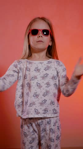 Vertical. Joyful young girl in red sunglasses and pajamas actively applauds with her hands and rejoices enthusiastically. Happy child in pajamas laughs and rejoices in victory.