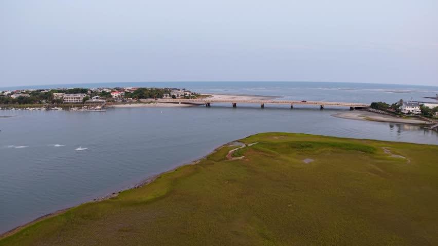 A drone soars over a tranquil marshland with a narrow causeway stretching across the water and distant houses lining the horizon.