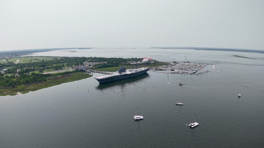 Drone video of the historic USS Yorktown aircraft carrier anchored in Charleston Harbor, with marshy wetlands, boats and a sprawling waterfront landscape under a clear sky.