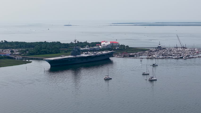 A high-altitude drone captures a wide shot of the USS Yorktown aircraft carrier docked at Patriots Point, surrounded by calm harbor waters and small boats.