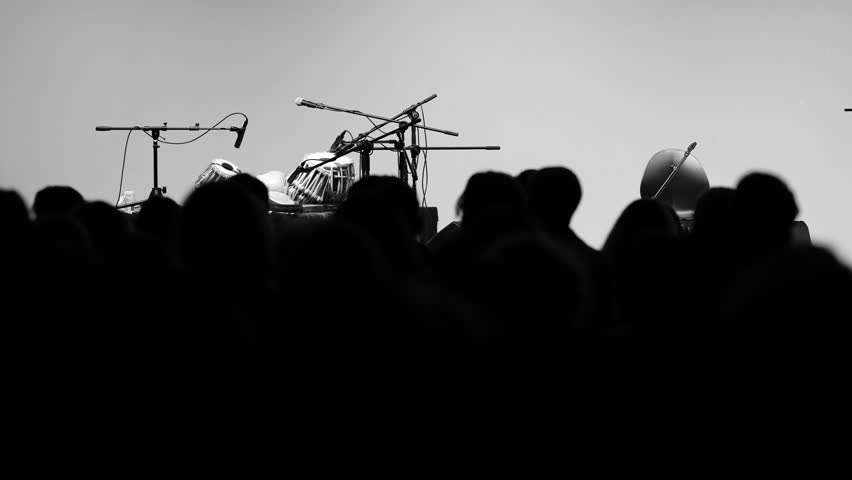 Black and white cinematic video. Silhouettes of the audience waiting for a concert. Microphones and Indian drums, like tabla, are prepared on stage. Moody atmospheric shadows and anticipation