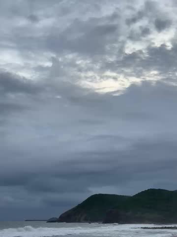 A dramatic coastal scene with dark storm clouds over the ocean at dusk. Waves crash onto the beach as a lone figure walks along the shore, creating a moody and atmospheric seascape.