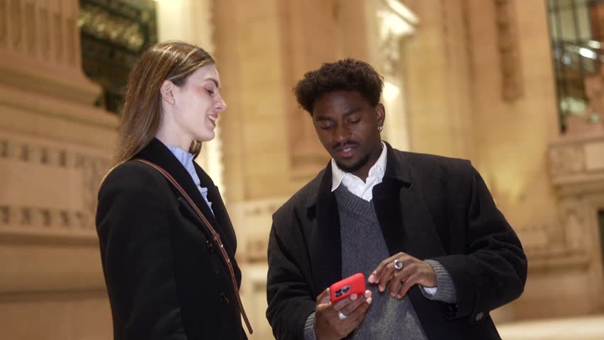 Happy young multiracial couple smiling and laughing while looking at a smartphone screen, enjoying a pleasant moment together outdoors at night in an urban setting with architectural background