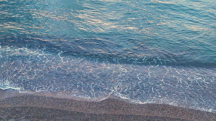 Aerial looking down shot of waves washing over colorful pebbles with rhythmic motion on the coast providing a serene and calming visual for nature documentaries and backgrounds