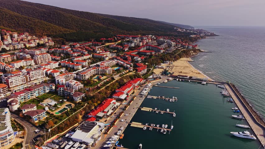 Aerial view captures a vibrant seaside resort in Bulgaria, showcasing colorful buildings, boats in the marina, and the beautiful coastline on a clear day.
