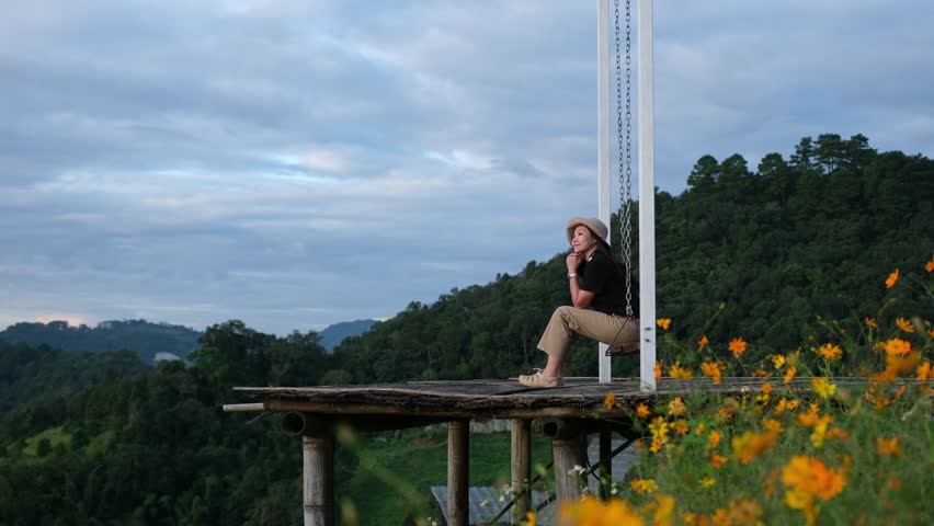 A woman sitting on a swing with a beautiful flower garden and mountain on cloudy day