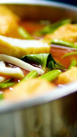 Close-up of spicy Asian red soup containing fried tofu, green vegetables, and fish cake in a metal bowl representing authentic delicious local cuisine