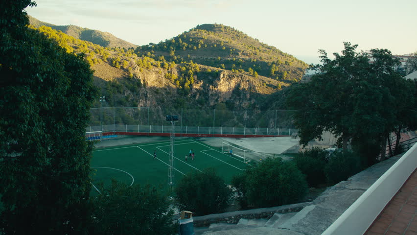 Children play football on a spectacular mountaintop field in southern Spain, surrounded by dramatic Andalusian cliffs and trees.