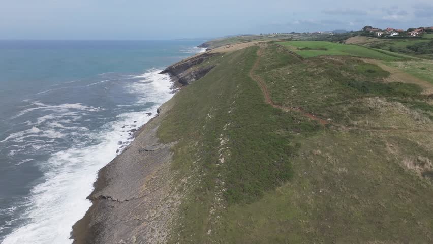A sideways flight leaves the green meadows and cliffs of the Cantabrian coast to fly over the open sea. A dividing line shows land on one side and water on the other; waves crash against the rocks.