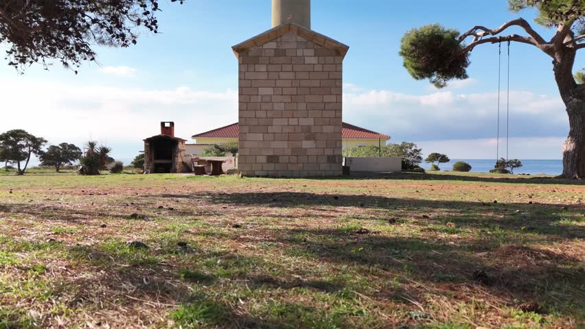Drone lifts through pine trees to reveal a small chapel and the Veli Rat lighthouse on Dugi Otok, with the blue Adriatic Sea in the background.