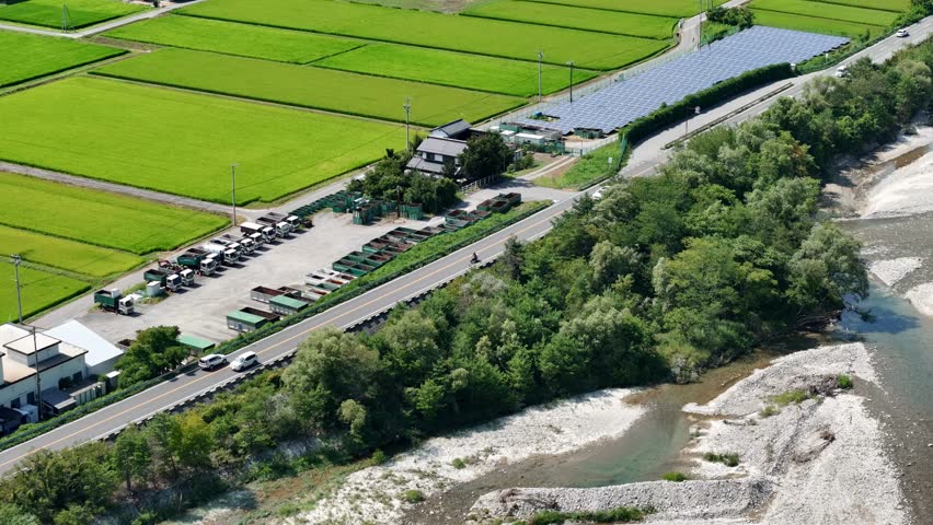 A view of a rural area with a lot of green rice fields and a road. The road is lined with trees and there are several cars parked along it
